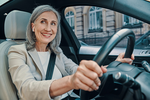 Mature beautiful woman in smart casual wear smiling and driving while sitting on front seat of the car