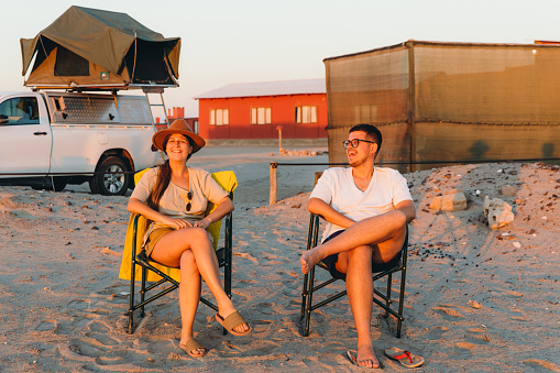 Young happy couple sitting at the beach camping with their 4X4 camper truck and looking at the sun going down in the ocean near Walvis Bay, Namibia