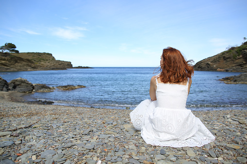 Back view of a woman in white dress sitting on the beach contemplating views