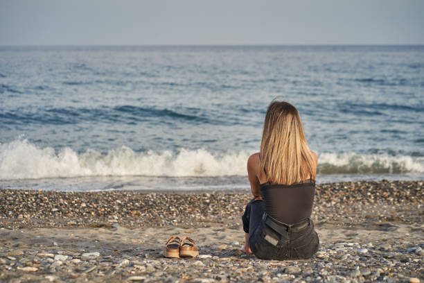 Woman sitting looking at the horizon with flip flops on the beach sand stock photo