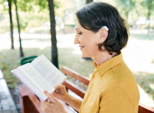mature woman with a hearing impairment uses a hearing aid in everyday life, reading a book in park, outdoor. hearing solutions - reading outside imagens e fotografias de stock