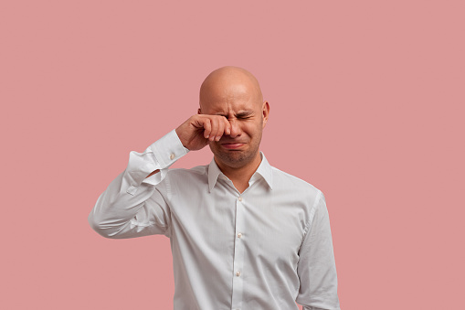 Unhappy sad bald man with bristle rubs tears wants to cry feels desperate has misunderstandings and bad conversation with people. Dressed in white shirt, isolated over pink background