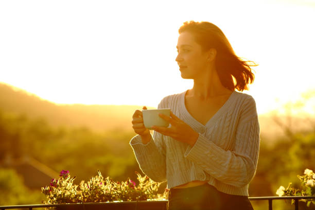 donna che tiene la tazza di caffè contemplando al tramonto in un balcone - balcony women patio coffee foto e immagini stock