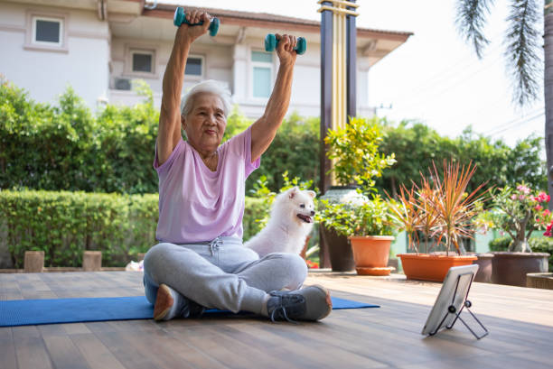 Home Workout Asian senior woman exercising with dumbbell while watching video from digital tablet at home. Health and technology. animal therapy stock pictures, royalty-free photos & images