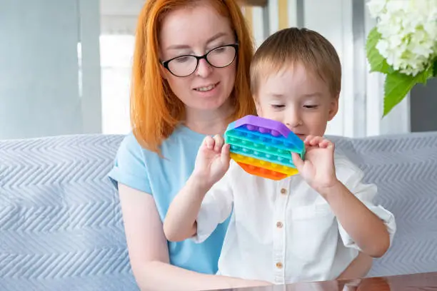 Photo of Mom and son are sitting on couch and playi with colorful fashion pop-it toy, an anti-stress sensitive toy or reusable bubble wrap. The trend of 2021.