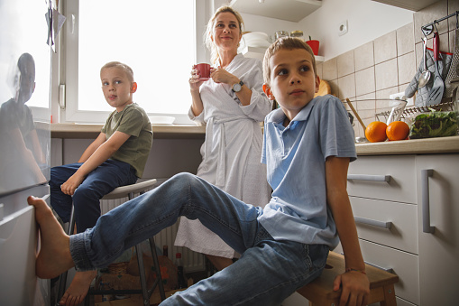 Low angle view of mid adult woman and her two sons looking away while having a quiet morning, drinking coffee, chatting and bonding in the kitchen.