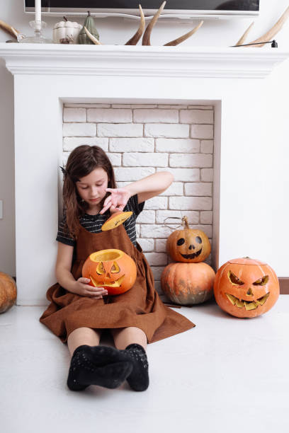 リビングルームの暖炉の隣に座って自宅でハロウィーンにカボチャを彫っている小さな女の子。お菓子をくれないといたずらするよ。ハロウィーンを祝う子供。 - six animals audio ストックフォトと画像