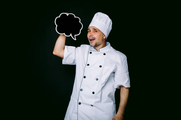 istock Chef cook wearing cooking jacket and hat, holding a blackboard with comic bubble speech, on black background 1327176868