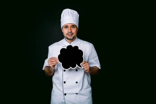istock Chef cook wearing cooking jacket and hat, holding a blackboard with comic bubble speech, on black background 1327176876