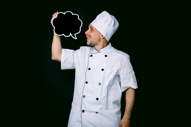 istock Chef cook wearing cooking jacket and hat, holding a blackboard with comic bubble speech, on black background 1327176899