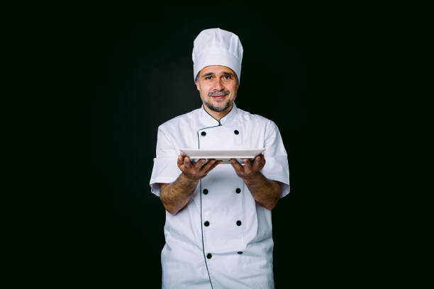 istock Chef cook wearing cooking jacket and hat, holding a plate, on black background 1327176910
