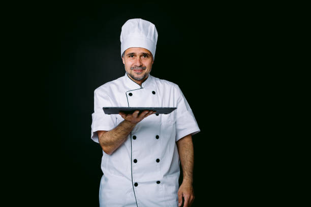 istock Chef cook wearing cooking jacket and hat, holding a slate plate, on black background 1327176918