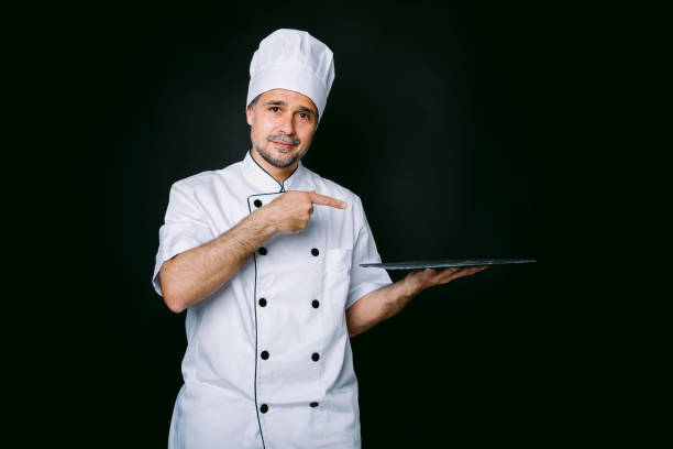 istock Chef cook wearing cooking jacket and hat, holding a slate plate, on black background 1327176931