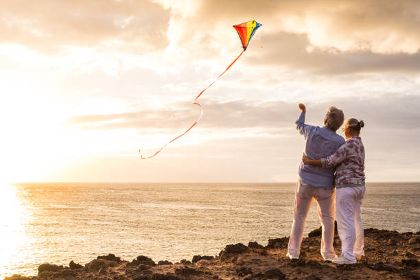 nahaufnahme und porträt von zwei alten und reifen menschen spielen und genießen mit einem flaying kite am strand mit dem meer im hintergrund mit sonnenuntergang - aktive senioren mit spaß - $89 stock-fotos und bilder