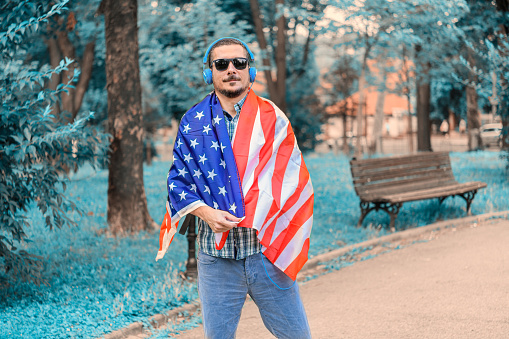 A Tourist With the Backpack is Relaxing With the Headphones on During a Walk Through the Public Park Full of Greeny Plants. A Man is Holding the US Flag in the Colorful Park.