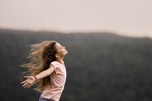 Happy little girl having fun with her arms outstretched and eyes closed during springtime in nature. Copy space.