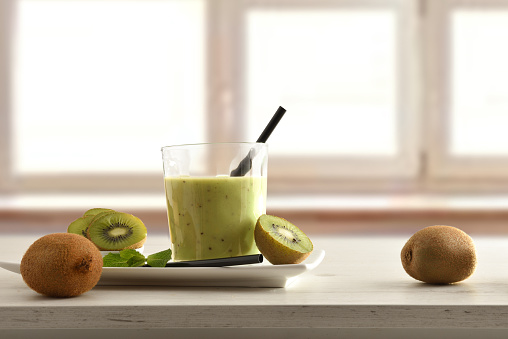 Fruit breakfast with glass of kiwi drink on plate and container cut fruit around on table and window background. Front view.