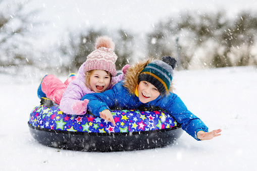 Active toddler girl and school boy sliding together down the hill on snow tube. Happy children, siblings having fun outdoors in winter on sledge. Brother and sister tubing snowy downhill, family time