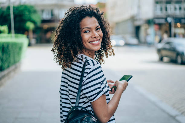Young smiling woman using smartphone on the street. Shot of beautiful young woman with smart phone texting outdoors in the city. looking over shoulder stock pictures, royalty-free photos & images