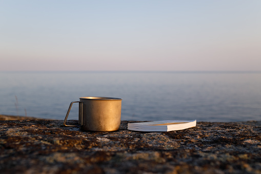 A mug with delicious tea and a book on the rock. A beautiful summer evening. The background of the sea is blurred.