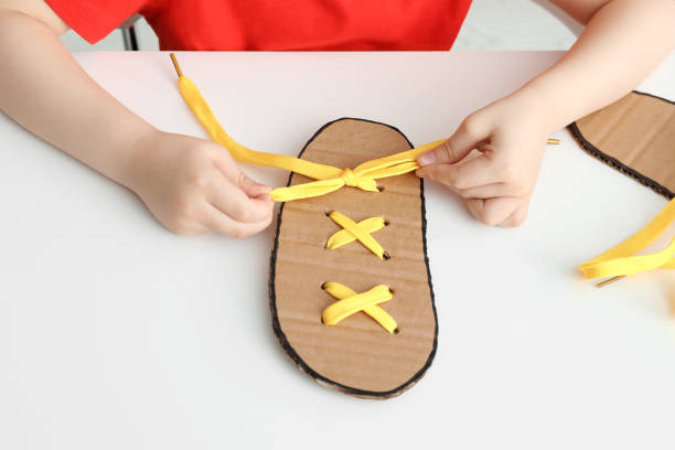 istock Little boy tying shoe lace using training cardboard template at white table, closeup 1327254276