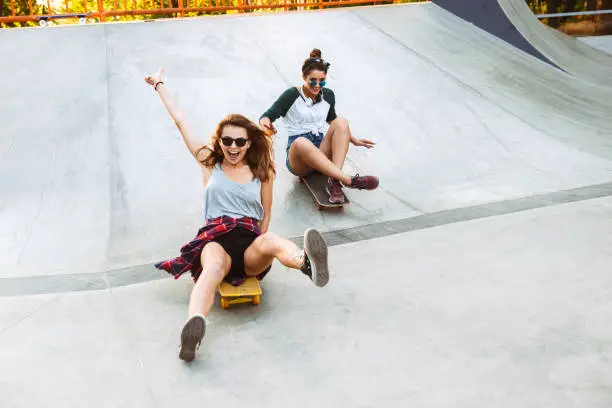 Two cheerful young girls having fun while riding on skateboards at the park