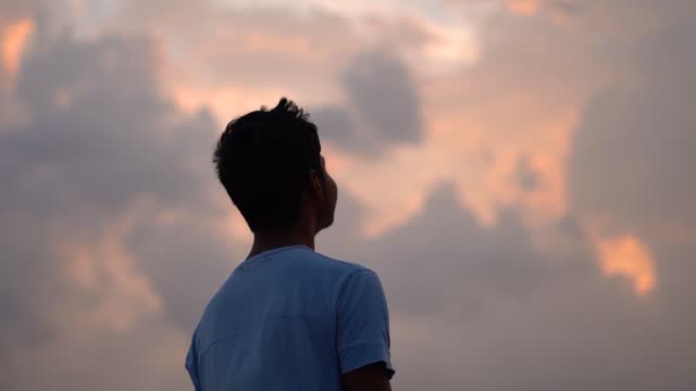 Silhouette of an Indian kid looking at the orange sky during the sunset. Hopeful kid stares at the sun. Kid lost in deep thoughts after looking at the sunset.
