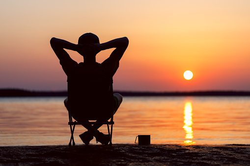A happy man is sitting on a camping chair with a mug of tea on the shore of a beautiful evening sunset sea. The background of the sea is blurred.