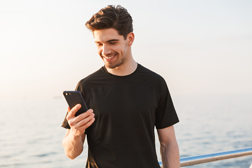 Smiling young sportsman in black t-shirt looking at mobile phone outdoors