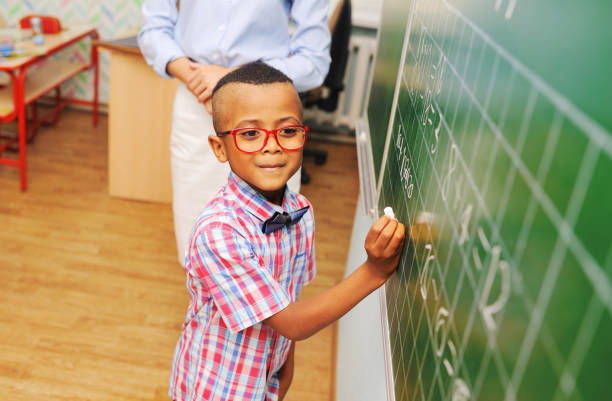 petit écolier afro-américain avec des lunettes écrivant à la craie sur le tableau noir contre l’enseignant - enfant prodige photos et images de collection