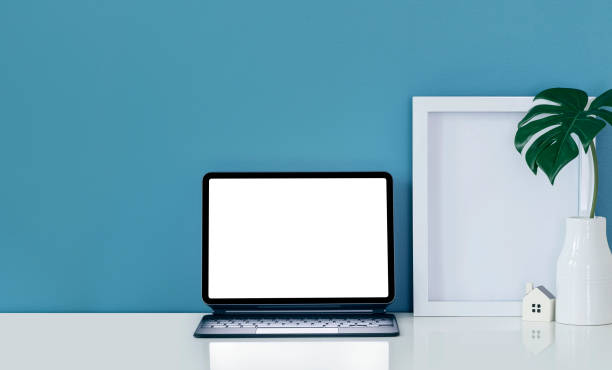 istock Tablet with magic keyboard, wooden frame and vase on white top table and blue wall. 1327260939