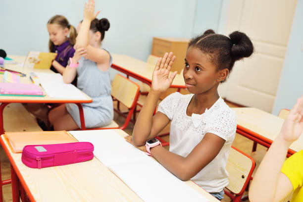 Small children school children smile and hold their hand up for a response against the background of a female teacher and a school class. Back to schoo a small African-American schoolgirl stretches her hand up and smiles against the background of a classroom and a group of classmates. Back to school, knowledge day. back to schoo stock pictures, royalty-free photos & images
