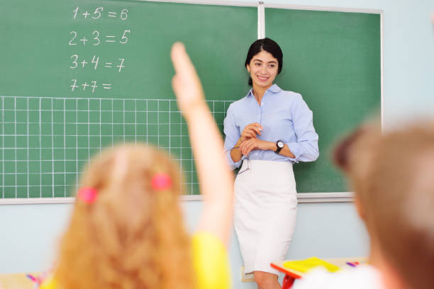 group of children sitting at their desks in a Junior elementary school hold out their hand for an answer against the background of a female teacher at the blackboard with mathematical examples. Back to schoo a group of children sitting at their desks in a Junior elementary school hold out their hand for an answer against the background of a female teacher at the blackboard with mathematical examples. Back to school, teacher's day, knowledge day back to schoo stock pictures, royalty-free photos & images