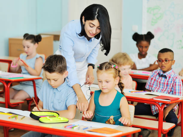 istock a young female teacher helps young children students with solving tasks against the background of a modern classroom in an elementary or primary school. Back to school 1327262563