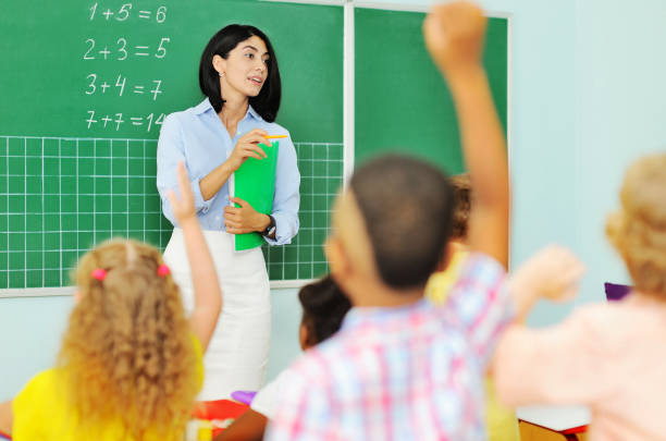 group of children sitting at their desks in a Junior elementary school hold out their hand for an answer against the background of a female teacher at the blackboard with mathematical examples. Back to schoo a group of children sitting at their desks in a Junior elementary school hold out their hand for an answer against the background of a female teacher at the blackboard with mathematical examples. Back to school, teacher's day, knowledge day back to schoo stock pictures, royalty-free photos & images
