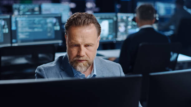 istock Portrait of Positive Professional Technical Controller Sitting at His Desk with Multiple Computer Displays Before Him. In the Background His Colleagues Working in System Control and Monitoring Center. 1327262795