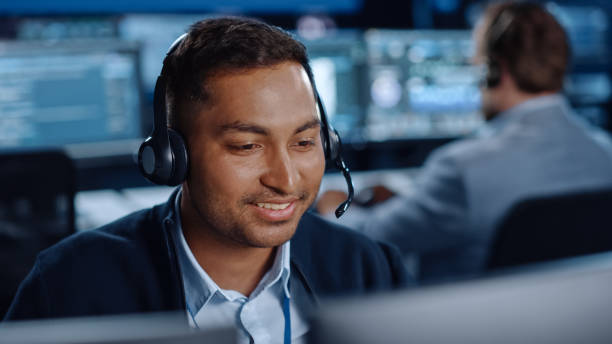 gros plan portrait d’un joyeux spécialiste du support technique à la clientèle parlant sur un casque tout en travaillant sur un ordinateur dans une salle de contrôle du centre d’appels remplie d’écrans d’affichage d’ordinateur et de serveurs - telephone network control room photos et images de collection