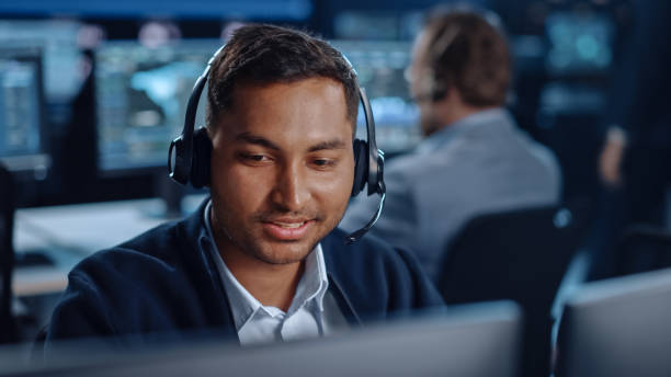 istock Close Up Portrait of a Joyful Technical Customer Support Specialist Talking on a Headset while Working on a Computer in a Call Center Control Room Filled with Computer Display Screens and Data Servers 1327262807