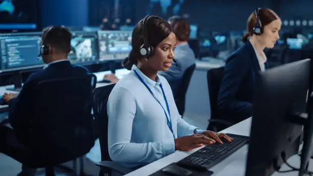 Photo of Happy Beautiful Technical Customer Support Specialist is Talking on a Headset while Working on a Computer in a Call Center Control Room Filled with Colleagues, Display Screens and Data Servers.