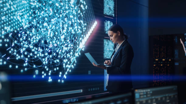 istock Young Female Computer Science Engineer Working on Laptop next at Big Screen Display Showing Neural Data. Telecommunications Technology Company System Control and Monitoring Room with Servers. 1327262861