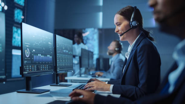 happy confident spécialiste du support technique à la clientèle ayant un appel de casque tout en travaillant sur un ordinateur dans une salle de surveillance et de contrôle sombre remplie de collègues et d’écrans d’affichage. - telephone network control room photos et images de collection