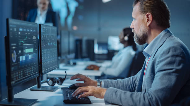 istock Portrait of Serious Professional Technical Controller Sitting at His Desk with Multiple Computer Displays Before Him. In the Background His Colleagues Working in System Control and Monitoring Center. 1327262881