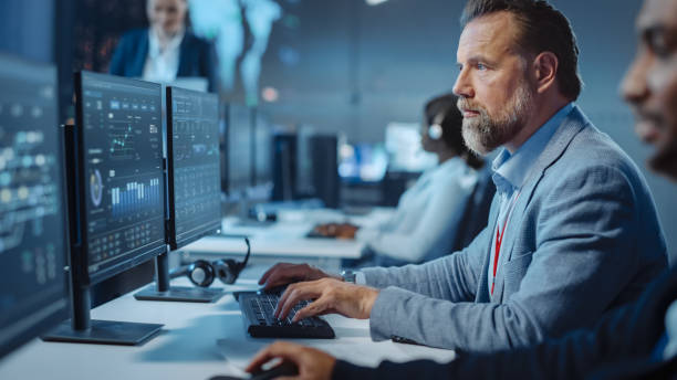 Portrait of Serious Professional Technical Controller Sitting at His Desk with Multiple Computer Displays Before Him. In the Background His Colleagues Working in System Control and Monitoring Center. Portrait of Serious Professional Technical Controller Sitting at His Desk with Multiple Computer Displays Before Him. In the Background His Colleagues Working in System Control and Monitoring Center. older software engineer stock pictures, royalty-free photos & images