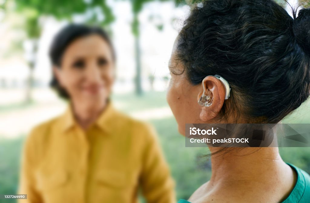 Adult woman with a hearing impairment uses a hearing aid to communicate with her female friend at city park. Hearing solutions Hearing Aid Stock Photo