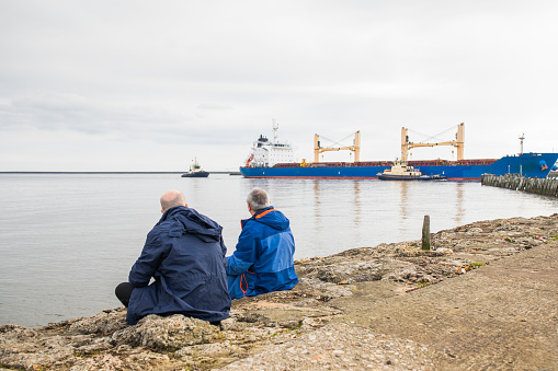 An over-the-shoulder view of two senior men sitting on the pier in North Shields in the North East of England, They are watching the ships sail into the marina.