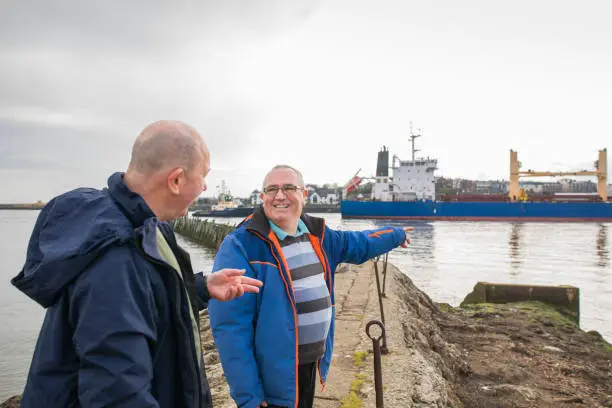 A side view of two senior men watching the big freight ships passing them from the Marina in North Shields Fish Quay. They are standing on the actual man-made pier and looking at each other.