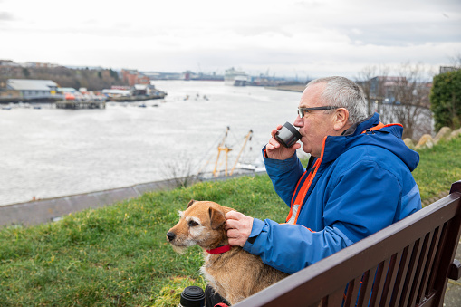 A side view of a senior man and his Patterdale Terrier dog sitting along the banks of the river tyne enjoying some quality time together on a dog walk in North Shields as they wait for a friend to join them.