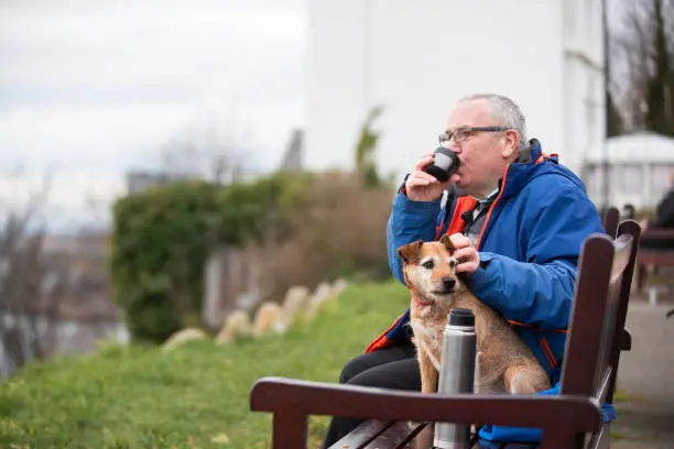 A side view of a senior man sitting on the banks of the river tyne with his pet Patterdale Terrier. He is sitting on a park bench and is looking out to the quayside and is having a hot drink on a fresh spring morning in North Shields.