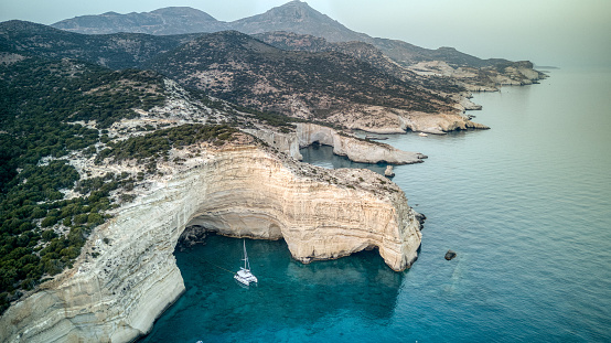 Aerial view of the dramatic Kleftiko Bay on Milos Island, Greece, with a sailing catamaran anchored in front of a sandstone cliff
