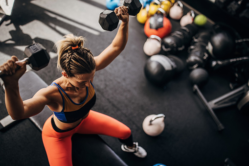 One woman, beautiful fit woman sitting on weight bench, exercising with dumbbells in gym.